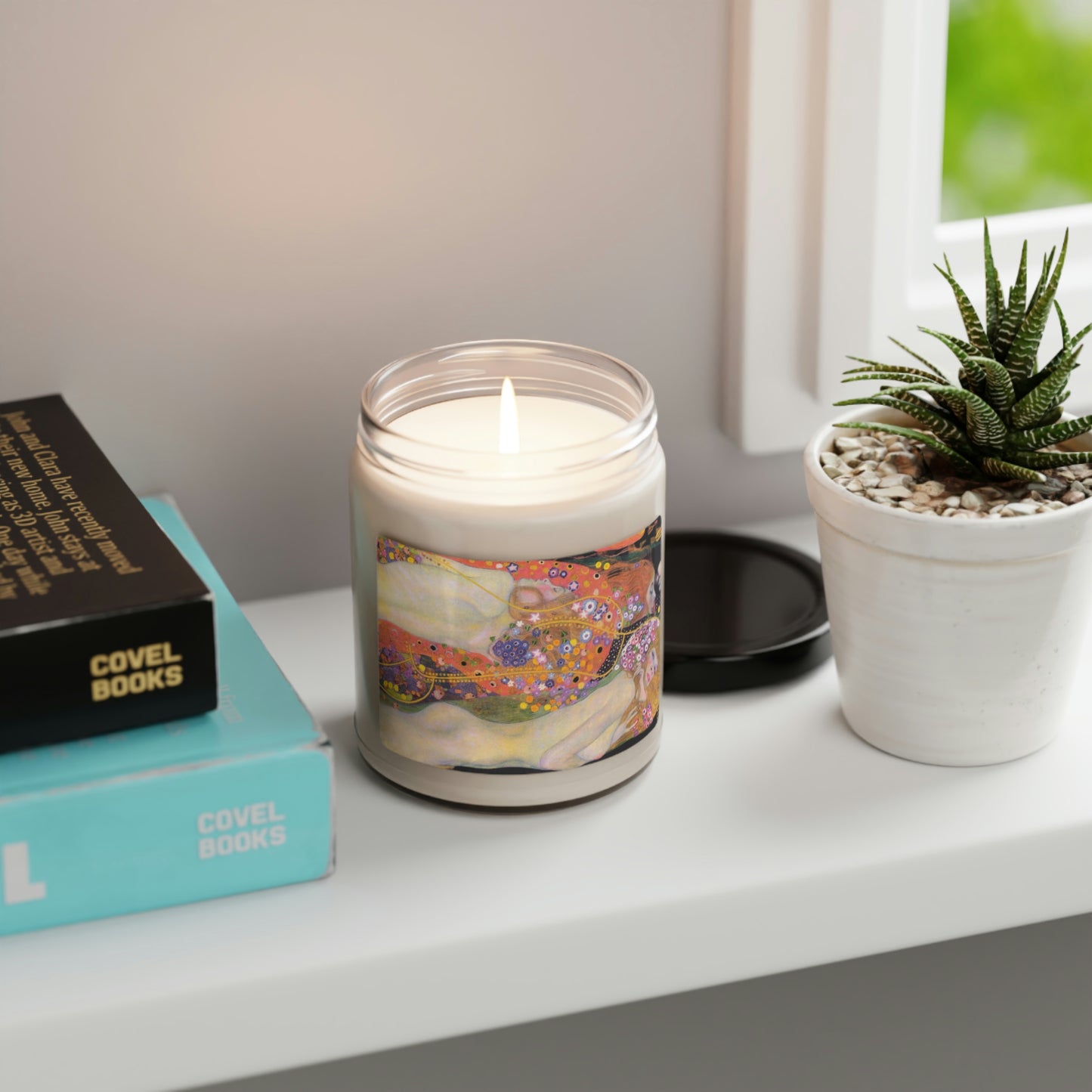 Scented Soy Candle of "Cinnamon Vanilla" with Klimt"Water Spirits' art, lit, on white table by aloe plant and book.