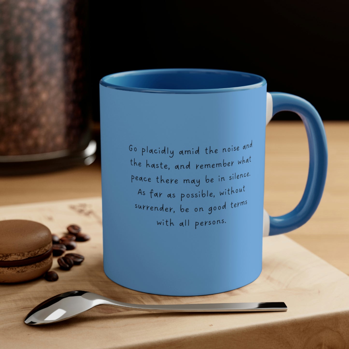 Light blue mug with bluer interior and handle, with quote "Desiderata. Go Placidly Among the Noise" sitting on a woodblock near a spoon and cookie