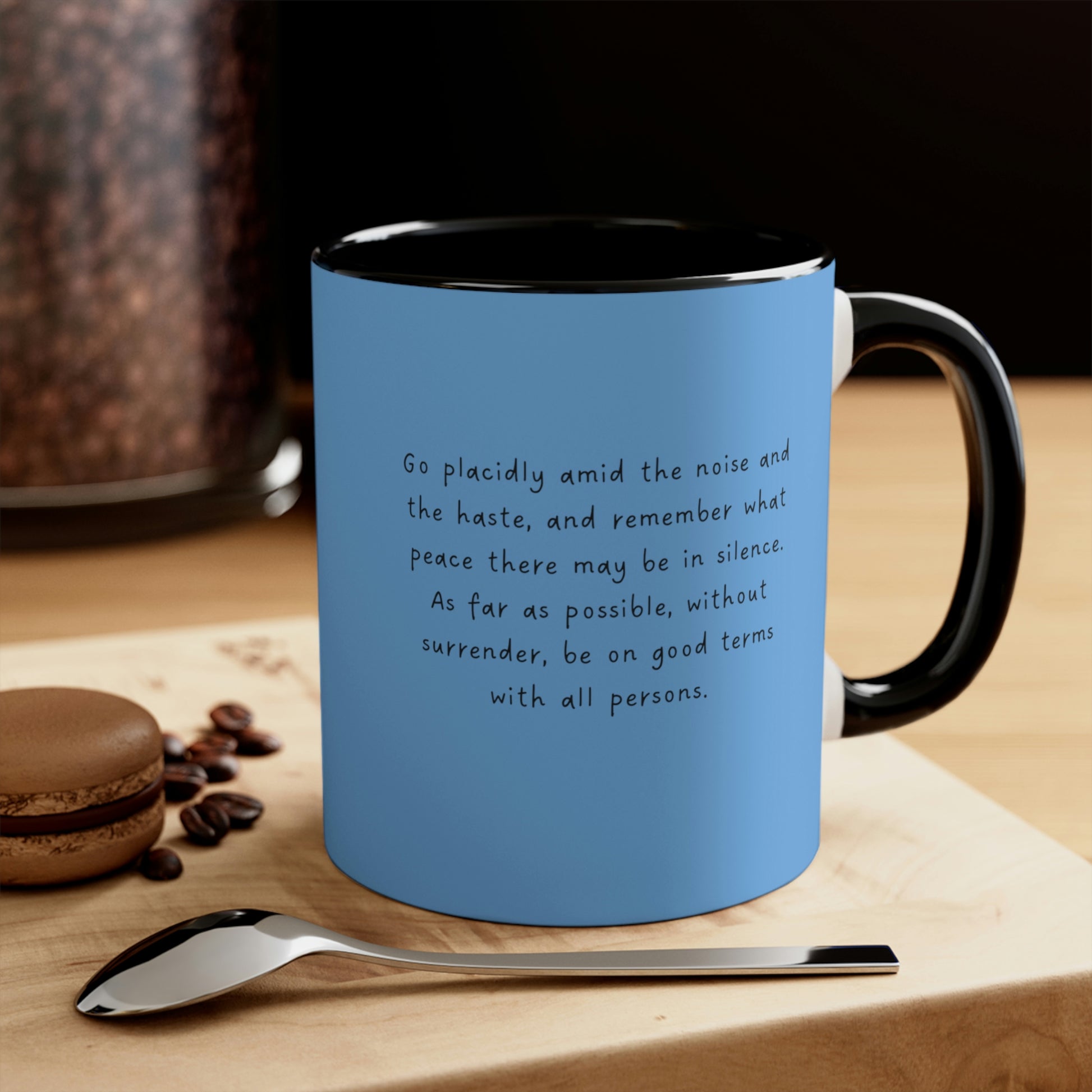 Light blue mug with black interior and handle with quote from Desiderata. "Go Placidly Among the Noise", sitting on a woodblock by a cookie and spoon.